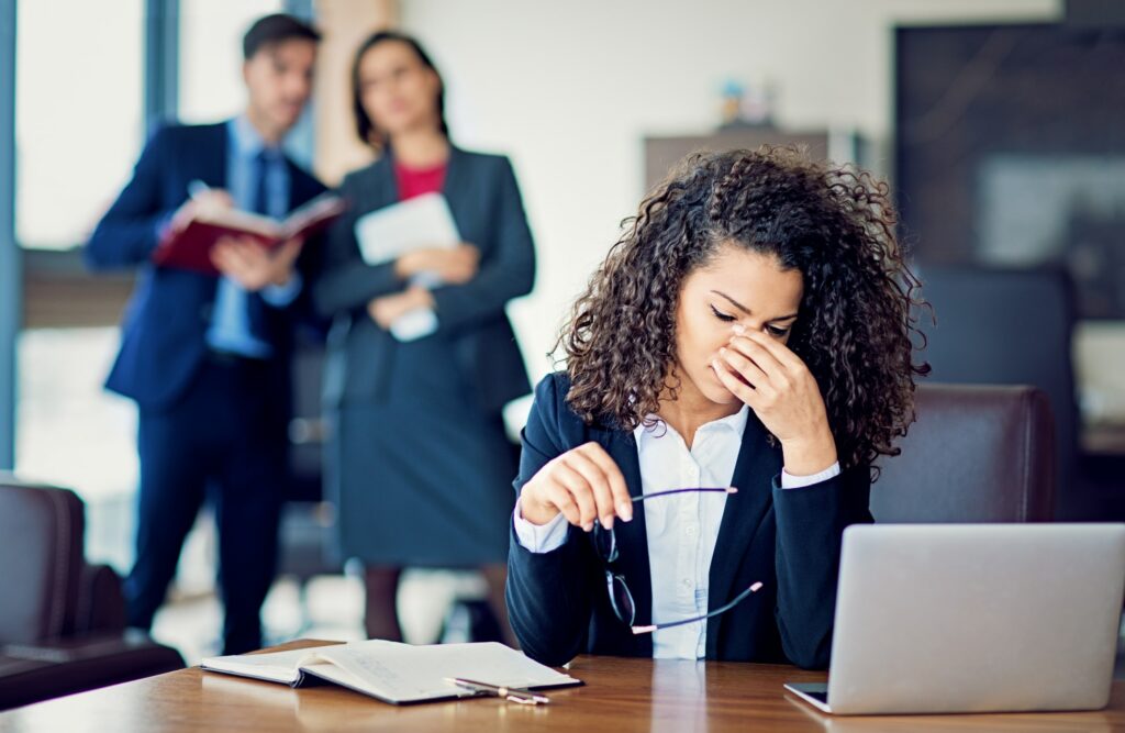 Discouraged employee sitting at desk after being bullied while other employees watch in the distance