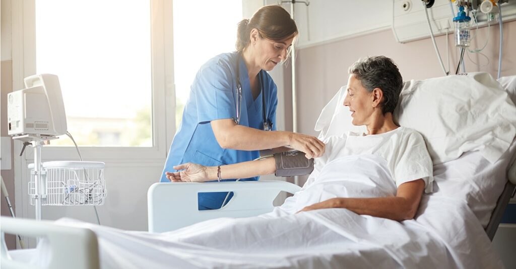 Female nurse leaning over bedside while attaching the blood pressure armband to a female patient's right arm.