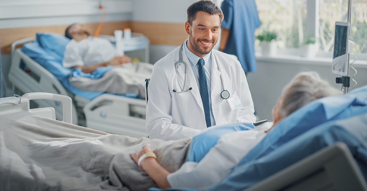 Smiling doctor in a white coat and stethoscope checks on a patient in a hospital bed.