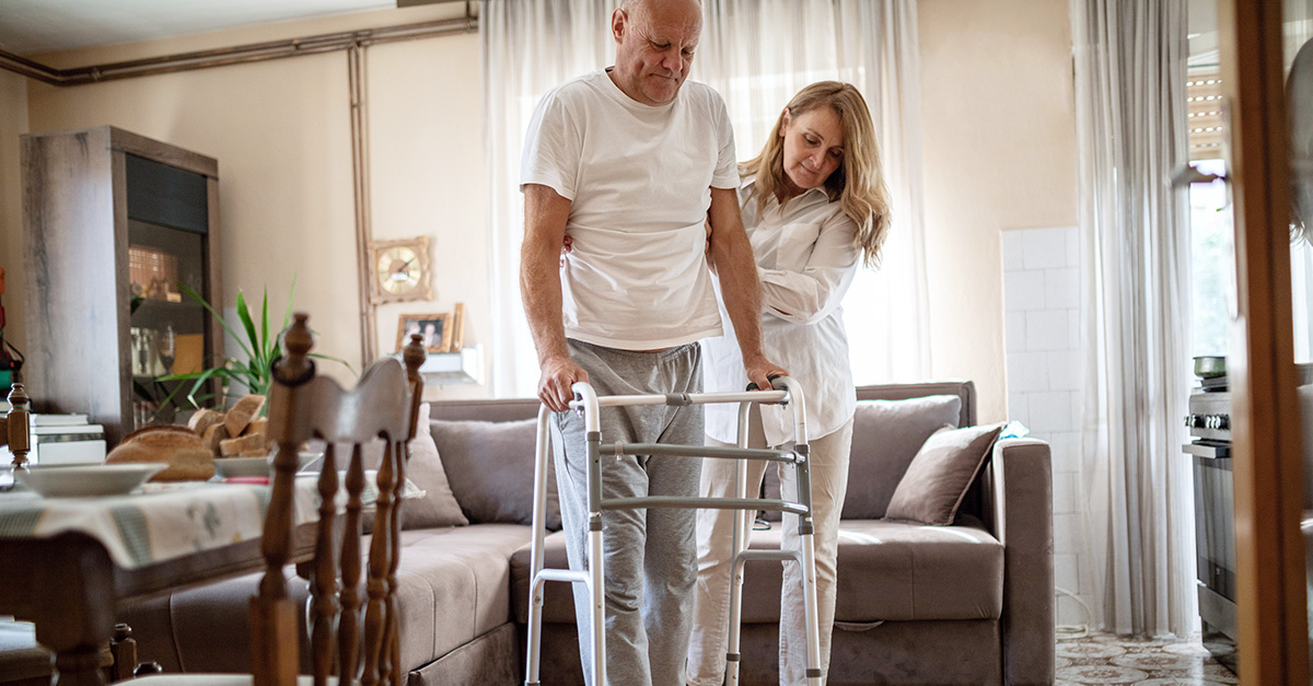 Wife helping her injured husband to walk using walking frame at home